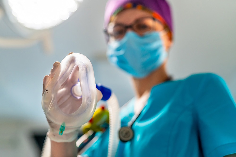 A healthcare worker in scrubs, mask, and gloves holds an anesthesia mask toward the camera, preparing for a medical procedure in a brightly lit clinical setting.