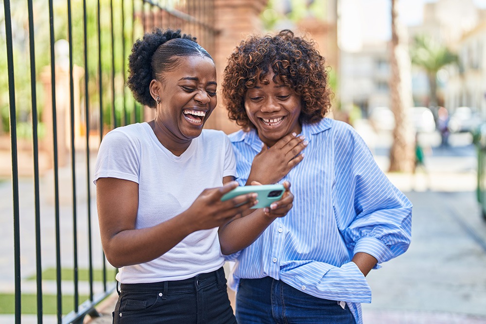 Two women standing outside, smiling and laughing while looking at a smartphone together. One wears a white t-shirt and holds the phone, the other wears a blue striped shirt with a hand on her chest.