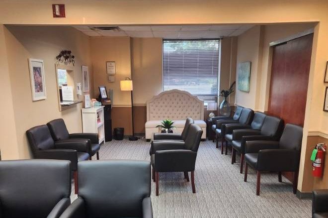 A waiting room with black chairs arranged along the walls, a white cushioned loveseat by a large window, a small plant in the center, and neutral-colored walls with framed artwork.