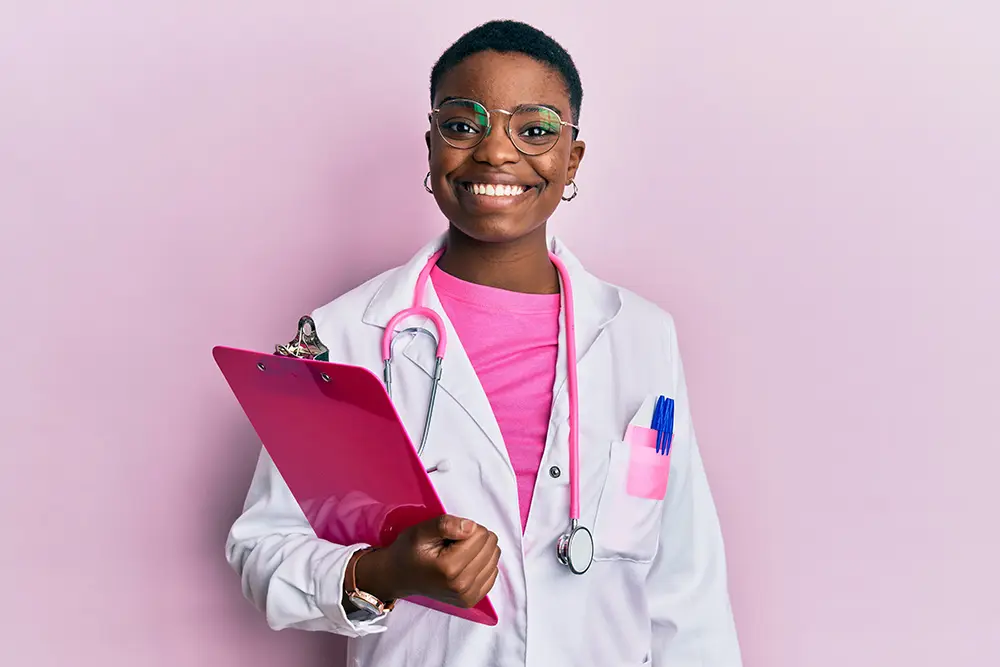 A smiling doctor with short hair and glasses wears a white coat and a pink shirt. She holds a pink clipboard and has a stethoscope around her neck, standing against a light pink background.