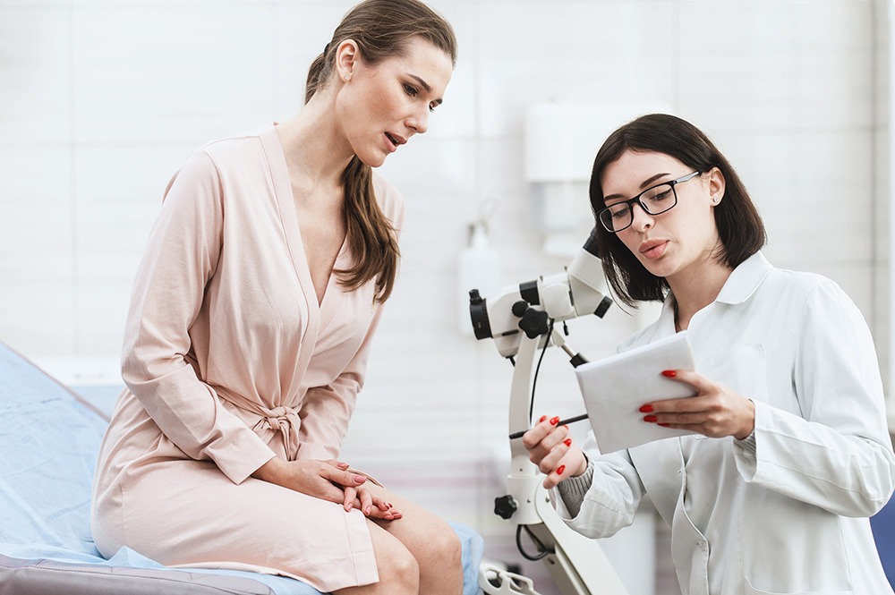 A doctor in a white coat shows a clipboard to a woman in a light pink medical gown who is sitting on an exam table in a clinic, discussing test results or a medical procedure.