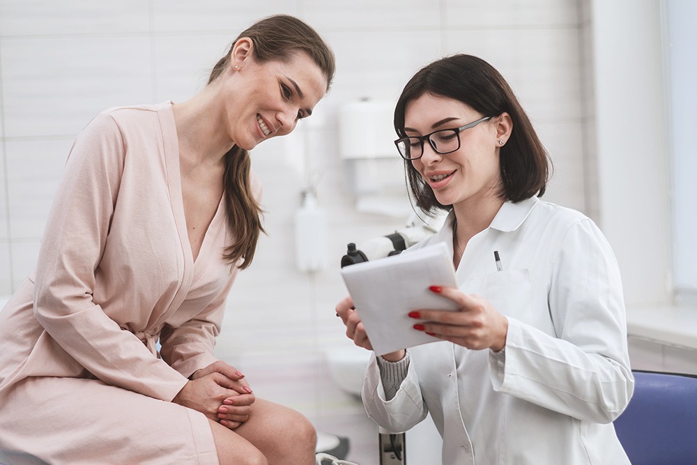 A female doctor wearing glasses holds a clipboard and discusses something with a smiling woman in a light robe, who is sitting in a medical office.