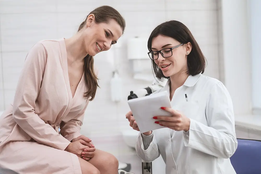 A female doctor wearing glasses holds a clipboard and discusses something with a smiling woman in a light robe, who is sitting in a medical office.