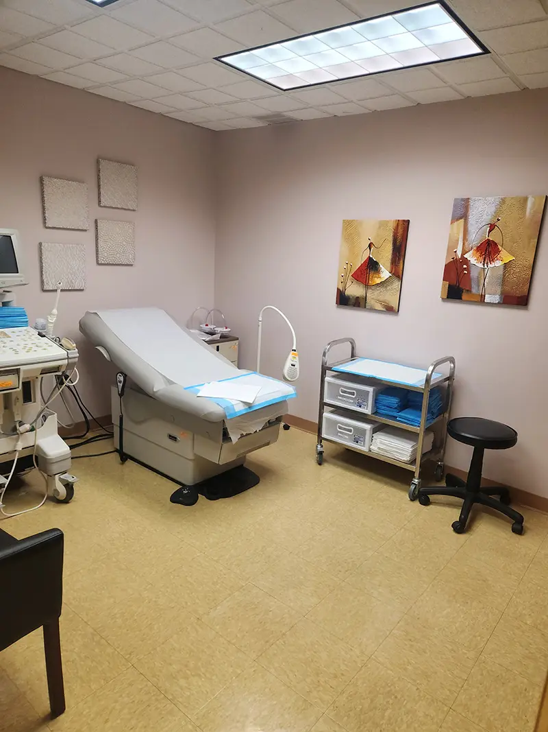 A clean medical exam room with an examination table, ultrasound machine, supply carts with folded linens, a stool, and two framed artworks on the wall. The room has beige walls and a bright overhead light.