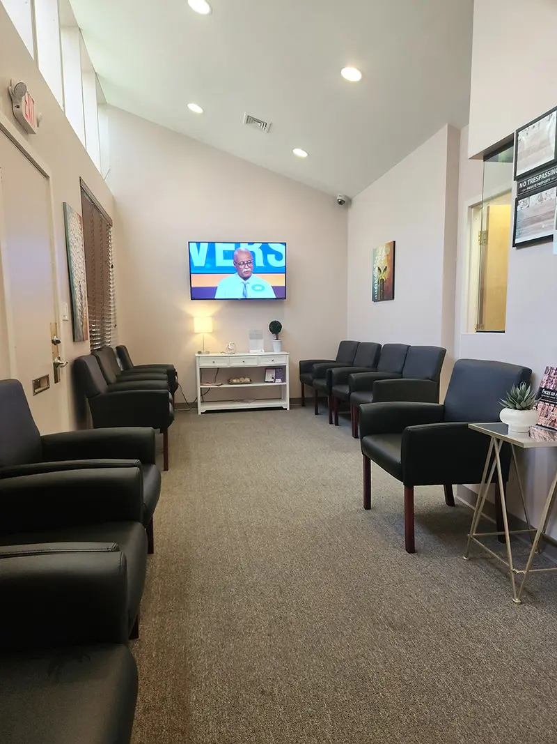 A clean, modern waiting room with black chairs lining both sides, a TV on the far wall displaying a man speaking, and neutral-colored walls and carpet. Brochures, artwork, and a small table are visible.