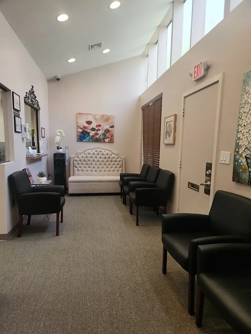 A waiting room with black chairs lined against the walls, a white upholstered bench at the back, wall art, a window, and an exit door under high windows letting in natural light.