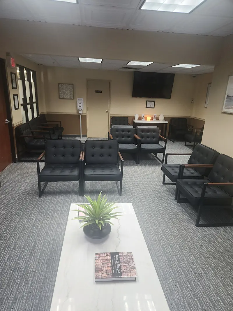 A waiting room with black cushioned chairs, a white coffee table with a plant and magazine, gray carpet, beige walls, a TV on the wall, and soft lighting. Hand sanitizer and decor are visible in the background.