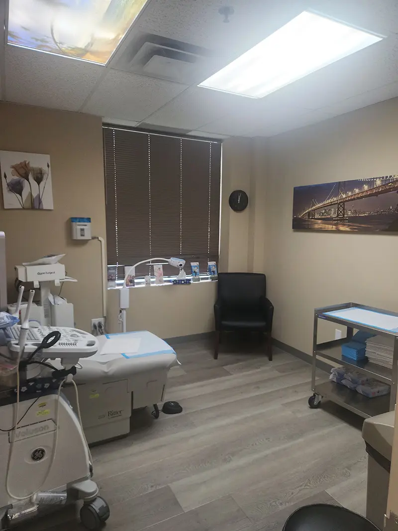 A clean medical examination room with equipment, an examination bed, a rolling cart with supplies, a black chair, wall art, and blinds covering the window; ceiling lights illuminate the space.