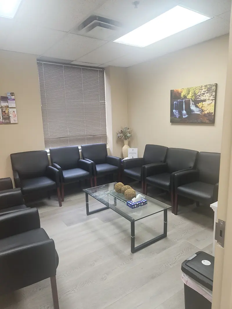 A waiting room with black chairs arranged along the walls, a glass coffee table with decorative items, a vase with flowers, a window with blinds, a wall painting, magazines, and a trash bin.