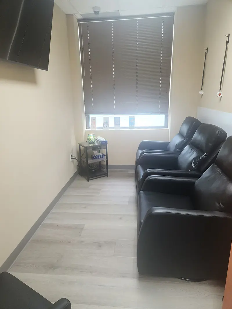 A small room with three black leather recliners, a window with closed blinds, a small shelf with items, hanging wall hooks, and a wall-mounted TV partially visible in the corner. Light wood flooring throughout.
