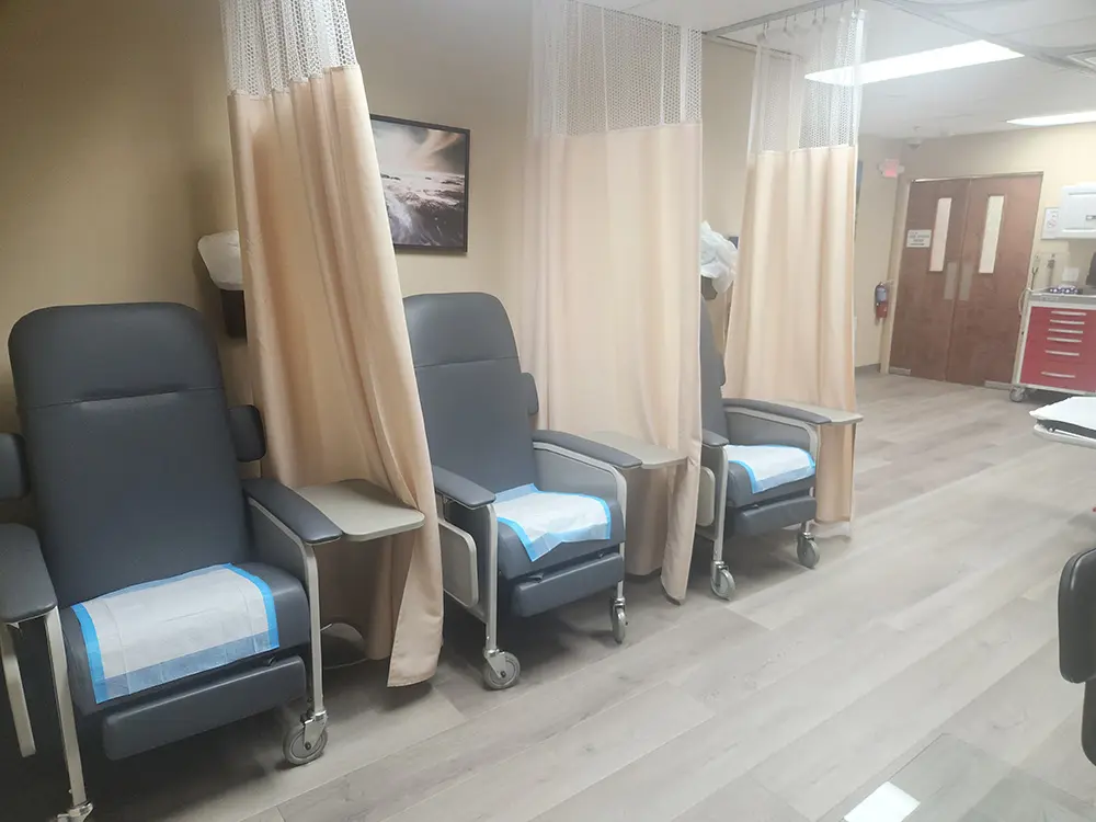 Three medical recliner chairs with blue pads are separated by beige privacy curtains in a clean clinic room with wood flooring, soft lighting, and medical equipment in the background.