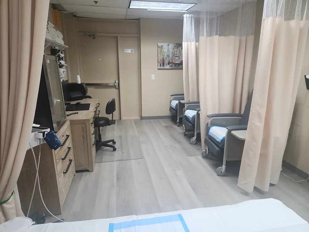 A medical clinic room with curtained patient areas, reclining chairs with blue pads, a workstation with a computer and chair, and medical equipment along the wall on light wood flooring.