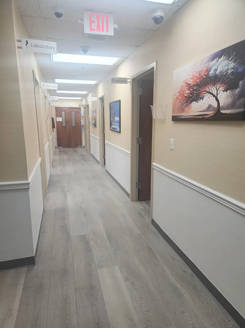 A clean, well-lit hallway in a medical office with light wood flooring, beige walls, framed artwork, and labeled doors including “Laboratory.” An exit sign hangs from the ceiling at the end of the hallway.