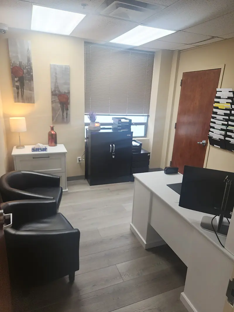 A small office with a white desk, computer, two black chairs, a file organizer, and a window with blinds. Wall art and a lamp decorate the beige walls, and a filing cabinet stands by the window.